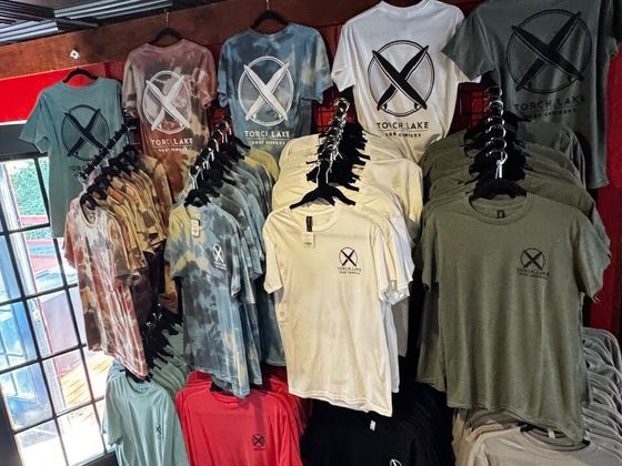A wall of Torch Lake Surf Company logo tees on hangers, in tie-dye, cream, olive and grey