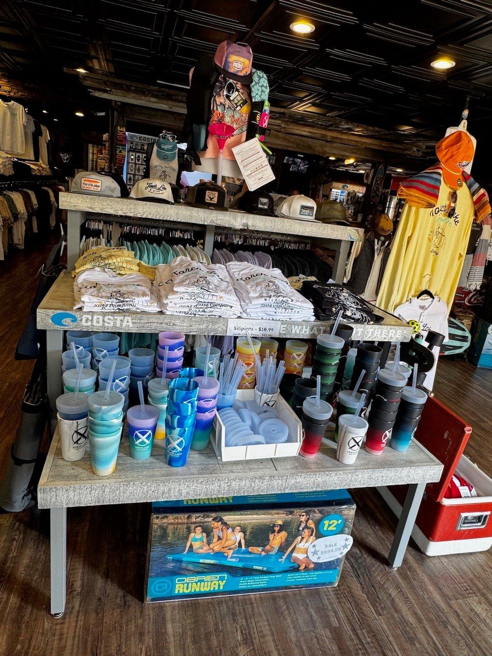 A center table of Silipint cups and insulated tumblers below folded tees and a shelf of caps