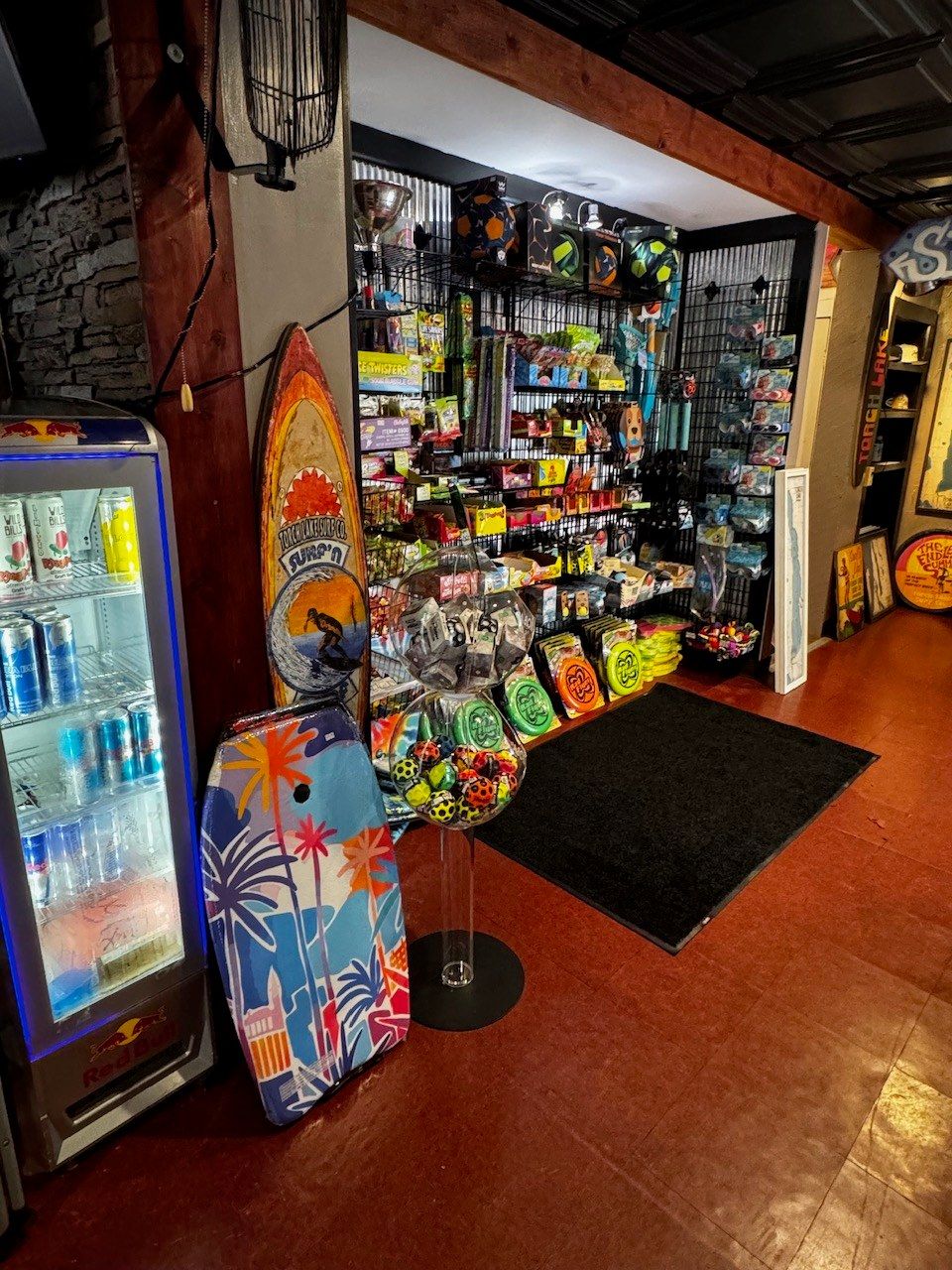 A drinks cooler and a vintage painted surfboard beside an alcove of candy, bodyboards and beach toys