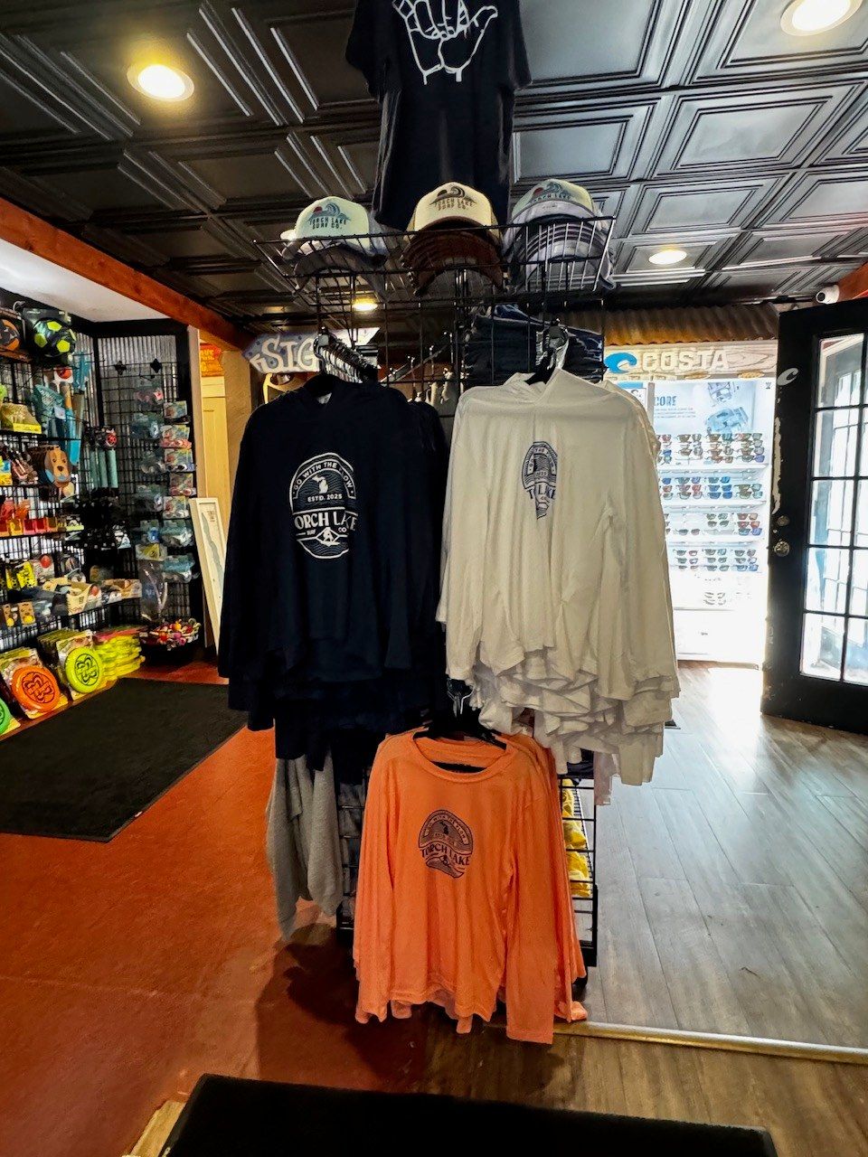 Long-sleeve tees on a spinner rack under a basket of caps, with the lit Costa sunglasses case and the front door behind