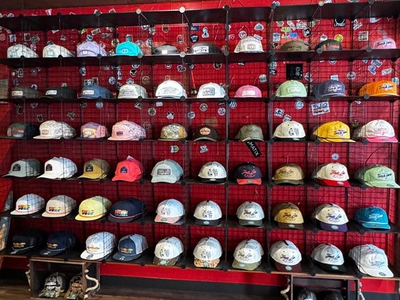 A red wall with a black wire grid holding dozens of caps, with patches and stickers pinned between them